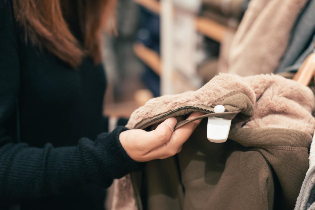 An image of a woman looking at a coat, inside a clothes store, with a security tag on it.